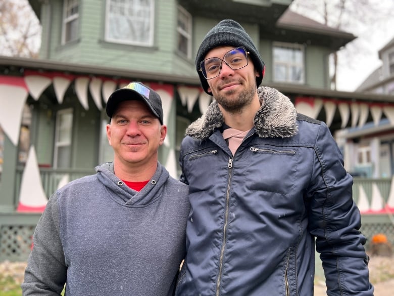 Two men stand in front of a house decorated with cardboard "fangs" along its front porch, making it look like a giant mouth.