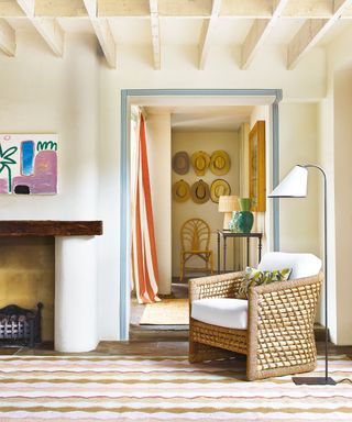 Cream living room with wooden ceiling beams, wicker armchair, sleek floor lamp, looking through to hallway, striped curtain, wooden console table with table lamp, colorful rug